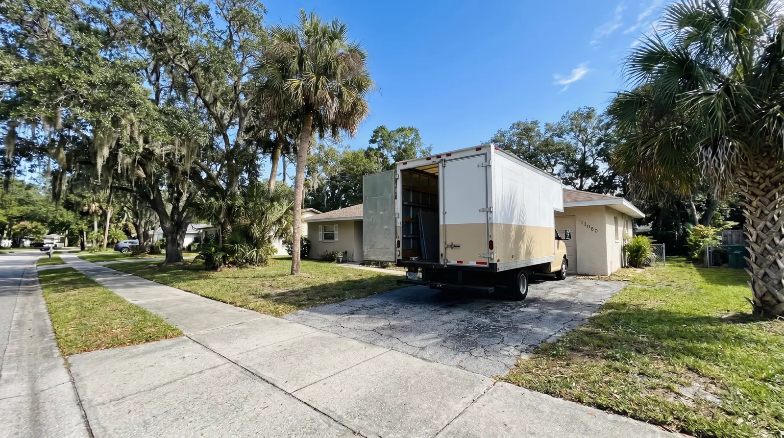 A moving truck parked in the driveway of a single-story ranch home on a quiet Spring Hill street lined with mature oaks and palmettos under a bright Florida sky